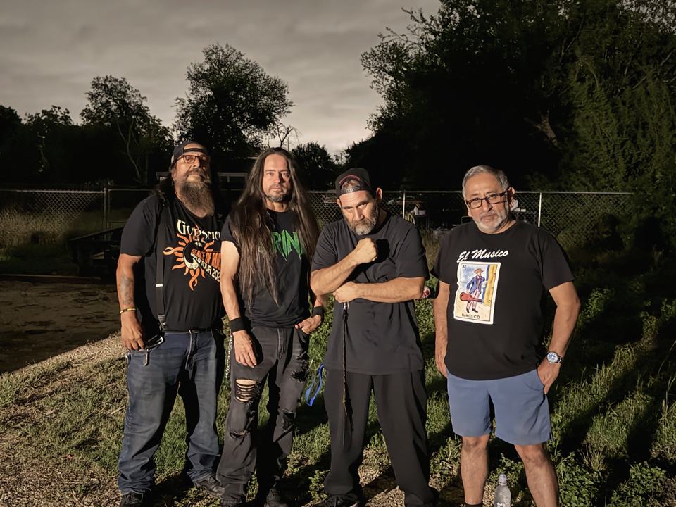 The four members of GRIND standing together outdoors at dusk in front of a chain-link fence and trees